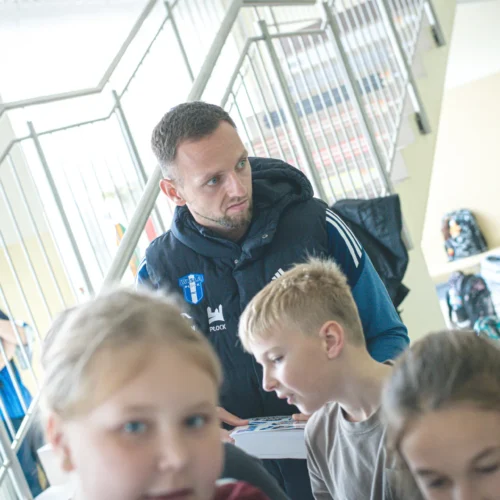 Na zdjęciu widać mężczyznę w ciemnej kamizelce, który rozmawia z grupą dzieci w szkolnym korytarzu.