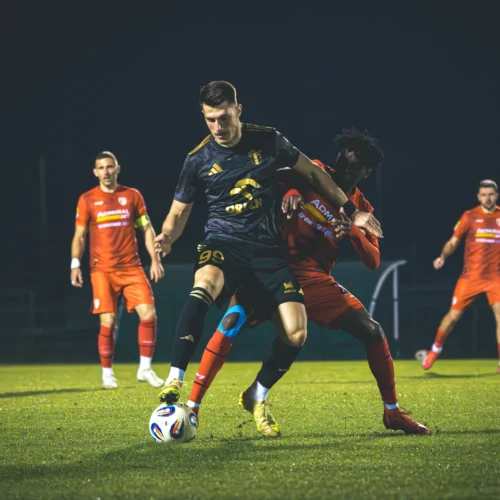 Na zdjęciu widać dynamiczną akcję meczu piłkarskiego, gdzie zawodnik w czarnym stroju walczy o piłkę z przeciwnikiem w czerwonym.