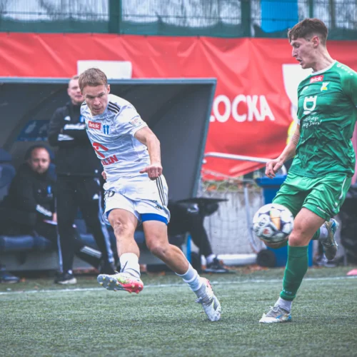 Na zdjęciu dwóch piłkarzy w akcji podczas meczu, jeden z zawodników w białej koszulce wykonuje strzał na bramkę, a drugi w zielonej koszulce jest w trakcie obrony.