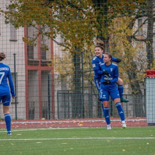 Na zdjęciu widoczne są radosne zawodniczki drużyny piłkarskiej w niebieskich strojach, celebrujące zdobycie gola na boisku.