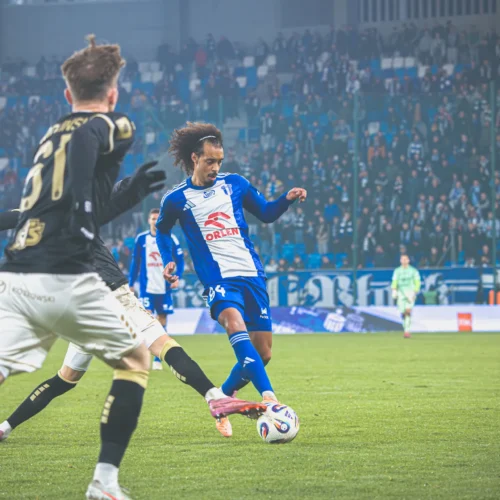 Na zdjęciu przedstawiony jest moment z meczu piłkarskiego, w którym zawodnik w niebieskim stroju wykonuje zagranie piłki, a w tle widać tłum kibiców.