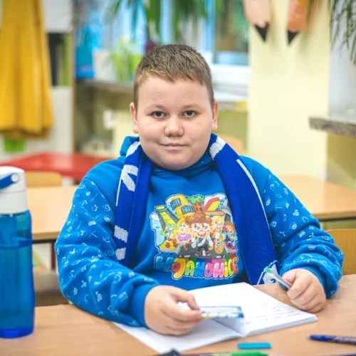 Chłopiec w niebieskiej bluzie z nadrukiem i szaliku siedzi przy biurku w klasie, trzymając długopis i notes.