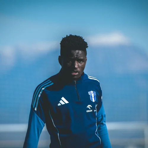 Mężczyzna w niebieskiej bluzie sportowej stoi na boisku piłkarskim, z wyraźnym skupieniem na twarzy.