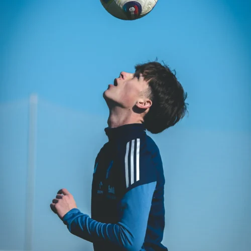 Chłopak z piłką nożną uniesioną nad głową stoi na boisku, wpatrując się w niebo.