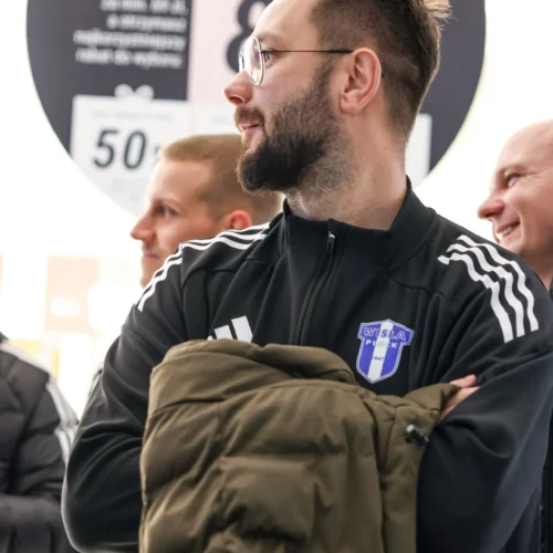 Mężczyzna w sportowej odzieży z logo Wisły Płock, trzymający kurtkę, stoi w towarzystwie innych osób w tle, na tle oznaczeń promocyjnych.