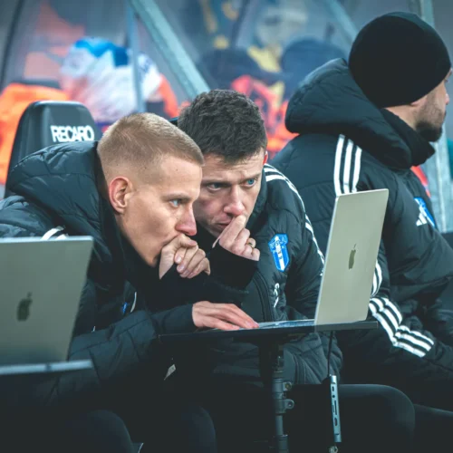 Na zdjęciu widać dwóch skupionych mężczyzn w czarnych kurtkach, siedzących na ławce rezerwowych, którzy uważnie przeglądają informacje na laptopach.