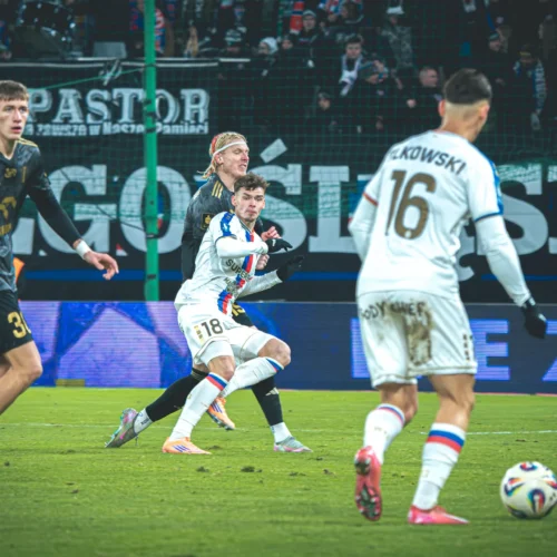 Na zdjęciu widoczni są piłkarze w akcji podczas meczu, z piłką na murawie i kibicami w tle.