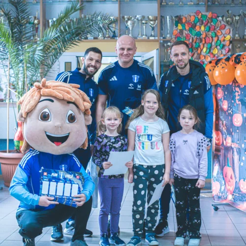 Na zdjęciu widać postać maskotki stojącą w towarzystwie trzech mężczyzn i czterech dzieci w radosnej atmosferze, z dekoracjami na Halloween w tle.