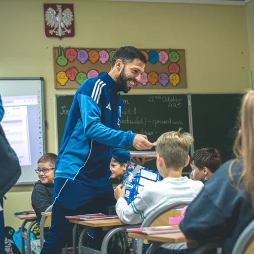 Na zdjęciu widać uśmiechniętego mężczyznę w niebieskim dresie, który rozmawia z dziećmi w klasie, a w tle znajdują się tablice edukacyjne.