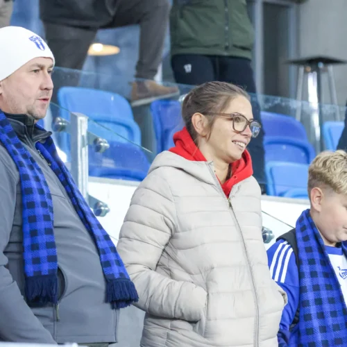 Na zdjęciu widać grupę osób, w tym mężczyznę, kobietę i chłopca, którzy są ubrani w ciepłe kurtki i szaliki, obserwujących wydarzenie sportowe na tle trybun.