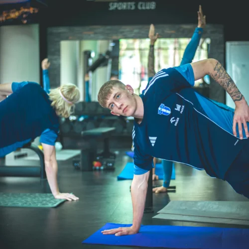 Na zdjęciu widać grupę osób wykonujących ćwiczenia w siłowni, w tym mężczyznę w niebieskim stroju, który trenuje na macie.