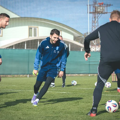 Na zdjęciu widać kilku piłkarzy trenujących na boisku, uśmiechających się i angażujących w ćwiczenia z piłką w słoneczny dzień.