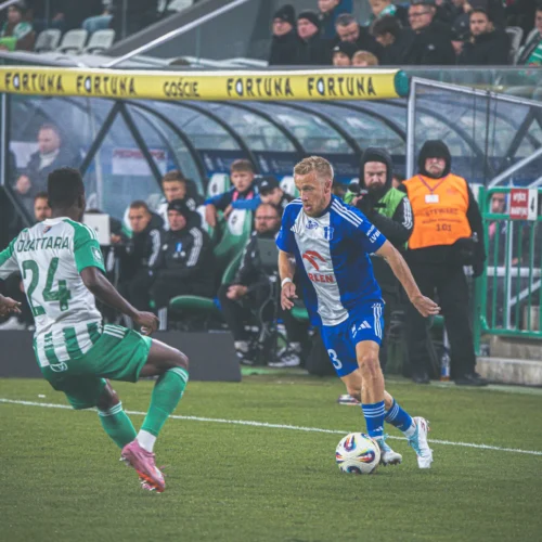 Na zdjęciu znajduje się dwóch piłkarzy w trakcie meczu, jeden z nich w niebieskim stroju atakuje, a drugi w zielono-białych barwach broni się na boisku.