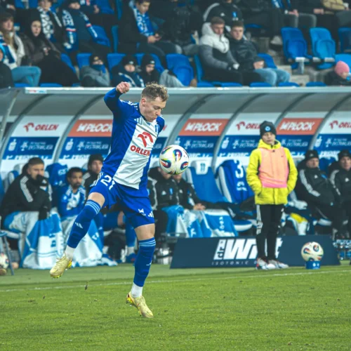 Na zdjęciu widać piłkarza skaczącego z piłką na boisku, z zasiadającymi w tle kibicami i ławką rezerwowych.