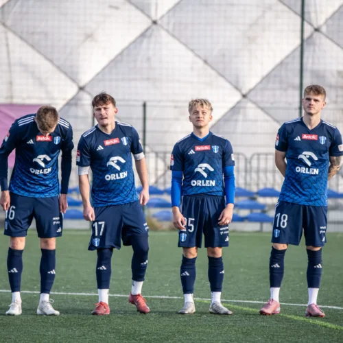 Na zdjęciu widać piłkarzy w niebieskich strojach meczowych, stojących na boisku przed rozpoczęciem gry.