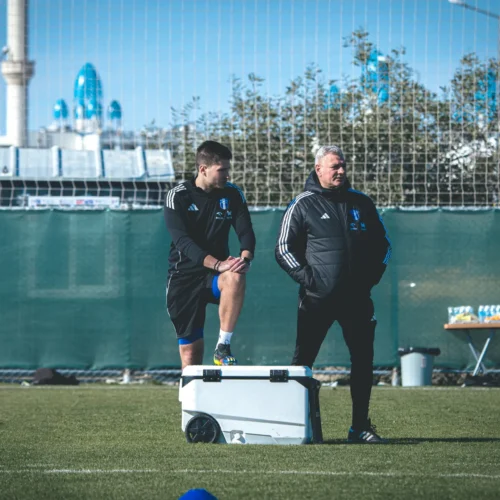 Na zdjęciu widoczny jest młody sportowiec stojący na skrzyni z uniesioną nogą oraz trener rozmawiający z nim podczas treningu piłkarskiego.