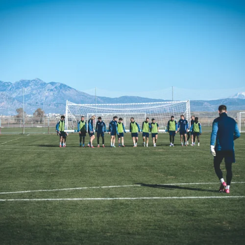 Na zdjęciu widać grupę piłkarzy w strojach treningowych, biorących udział w treningu na boisku z górskim tłem.