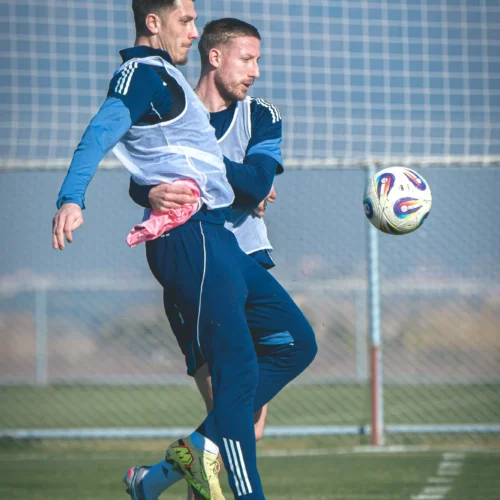 Dwaj piłkarze w niebieskich strojach trenują na boisku, jeden z nich podskakuje, próbując dosięgnąć piłkę.