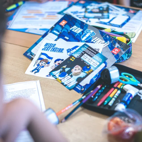 Na zdjęciu widać stół z kolorowymi materiałami do rysowania oraz kartkami związanymi z tematem sportowym, na których znajdują się grafiki postaci w strojach sportowych.