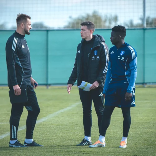 Na zdjęciu trzech mężczyzn rozmawia na boisku treningowym, omawiając strategię lub trening.