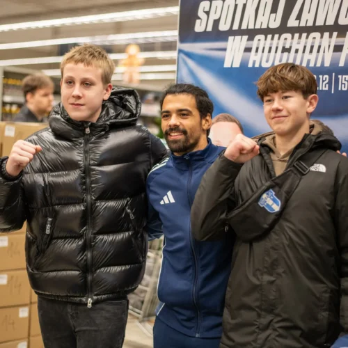 Dwóch młodych chłopców, w kurtkach puchowych, pozują z uśmiechem obok mężczyzny w granatowym dresie, na tle plakatu zapraszającego na spotkanie z zawodnikami w Auchan Płock.