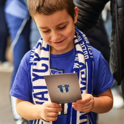 Chłopiec w niebieskiej koszulce i szaliku trzyma kartkę, na której znajduje się logo drużyny sportowej, z uśmiechem wpatrując się w nią wśród innych ludzi.