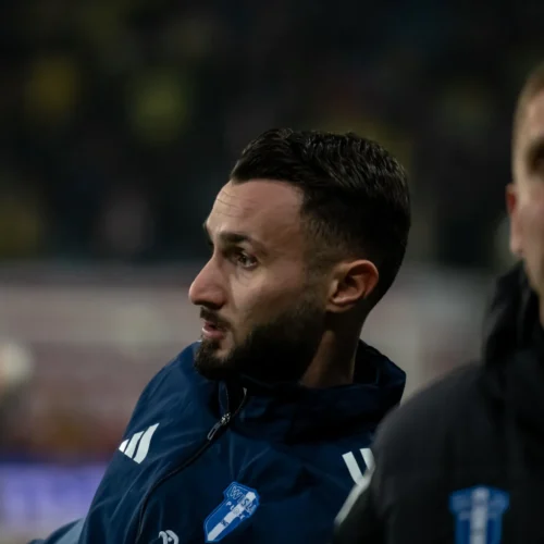 Na zdjęciu znajduje się mężczyzna w dresie sportowym, który wydaje się być skupiony na wydarzeniach na boisku, obok niego inny człowiek w ciemnej kurtce.