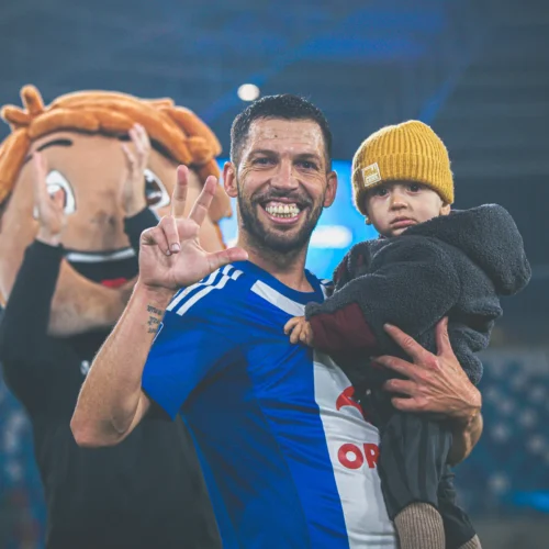 Na zdjęciu widoczny jest uśmiechnięty mężczyzna w niebieskiej koszulce sportowej trzymający małego chłopca, podczas gdy za nimi znajduje się postać maskotki.