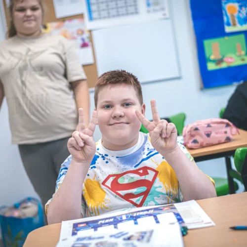 Na zdjęciu widać uśmiechniętego chłopca w koszulce z motywem superbohatera, który pokazuje znak pokoju, w towarzystwie innych dzieci w klasie.
