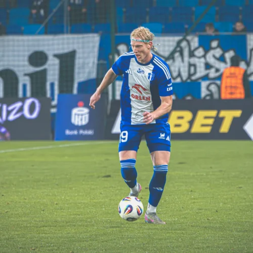 Na zdjęciu przedstawiony jest piłkarz w niebiesko-białym stroju, kontrolujący piłkę na boisku, z kibicami w tle.