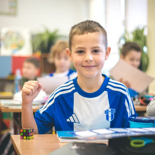 Chłopiec w niebieskiej koszulce sportowej uśmiecha się i pokazuje zaciśniętą pięść, siedząc przy biurku w klasie.