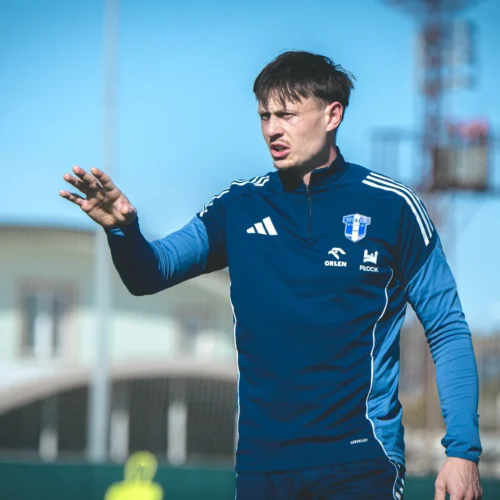 Mężczyzna w niebieskiej bluzie sportowej sygnowanej logotypami, gestykuluje w trakcie treningu piłkarskiego na zewnątrz w słoneczny dzień.