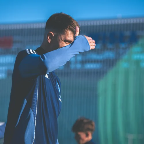 Na zdjęciu widać sportowca w niebieskim stroju, który zasłania oczy ręką, stojącego na treningu w słoneczny dzień.