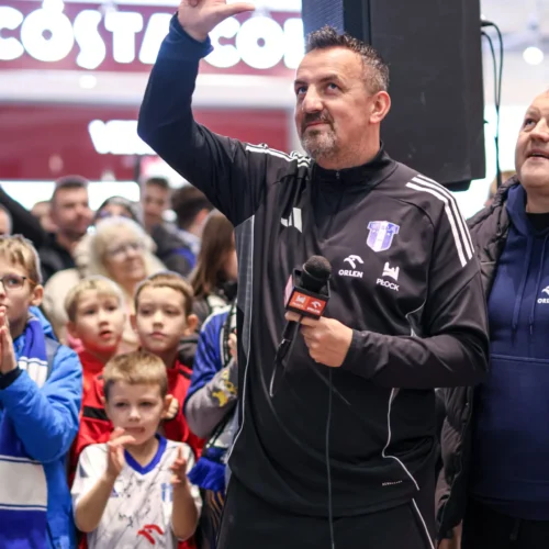 Mężczyzna w czarnym dresie stoi na scenie z mikrofonem, prezentując znak V, a w tle widać grupę zafascynowanych dzieci.