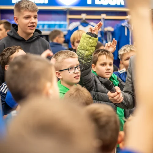 Grupa dzieci, z jednym chłopcem w zielonym swetrze, uczestniczy w wydarzeniu, trzymając ręce w górze i z zainteresowaniem słuchając dorosłego prowadzącego.