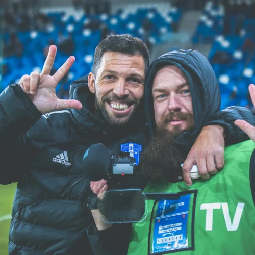 Dwie uśmiechnięte osoby na stadionie pozują do zdjęcia, trzymając gesty "V" i trzymając kamerę.