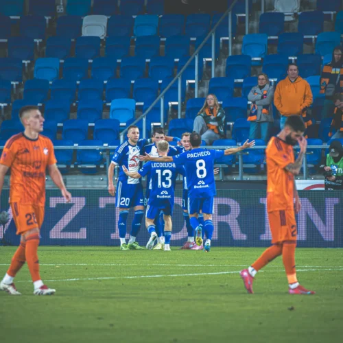 Na zdjęciu widoczni są piłkarze świętujący zdobycie gola, podczas meczu na stadionie, w tle widać kibiców w pomarańczowych koszulkach.