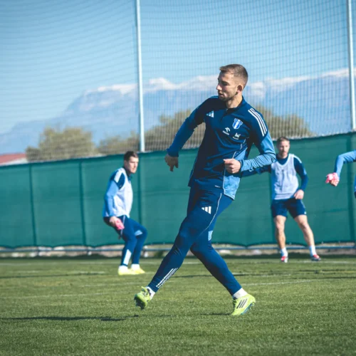 Na zdjęciu widać piłkarzy trenujących na boisku, w tle pasmo górskie.