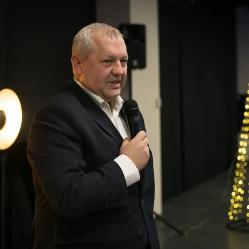 Mężczyzna w garniturze trzyma mikrofon i wygłasza przemówienie, a w tle widać dekoracyjnie oświetloną choinkę.