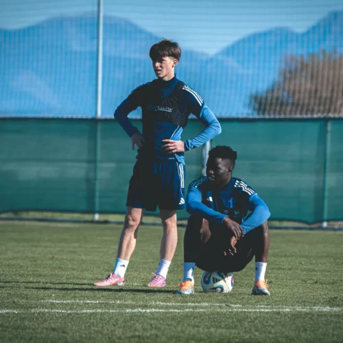 Dwaj młodzi piłkarze stoją i siedzą na boisku treningowym, z tłem gór i siatkami.