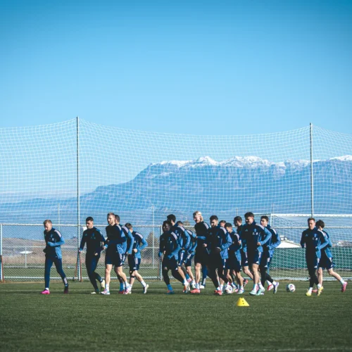 Na zdjęciu widać zespół piłkarski podczas treningu na zielonym boisku, z ośnieżonymi górami w tle.