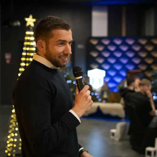 Mężczyzna w ciemnym swetrze trzyma mikrofon i uśmiecha się, prowadząc wydarzenie w eleganckiej sali udekorowanej choinką i oświetleniem.