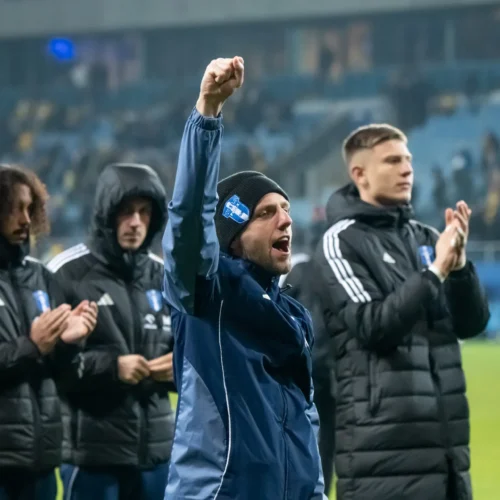 Na zdjęciu widoczna jest grupa mężczyzn w sportowych strojach, z jednym z nich podnoszącym rękę w geście radości na stadionie.