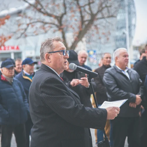 Mężczyzna w garniturze mówi do mikrofonu przed grupą słuchaczy na zewnątrz, w tle widoczne drzewa i budynki.