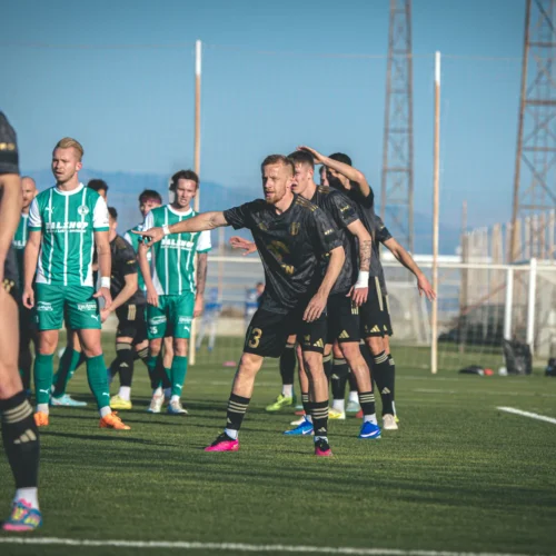 Na zdjęciu widoczna jest grupa piłkarzy w trakcie meczu, z których część ubiera się w czarne stroje, a inni w zielono-białe koszulki, na boisku otoczonym siatkami.