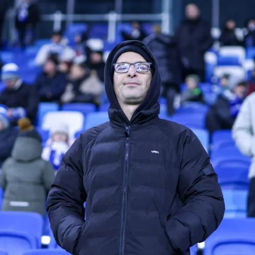 Mężczyzna w czarnej kurtce z kapturem stoi na stadionie, w otoczeniu innych widzów na trybunach.
