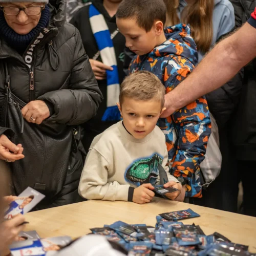 Na zdjęciu widoczna jest grupa ludzi, w tym młody chłopiec trzymający karty, z wyraźnym zainteresowaniem patrzący na otoczenie.