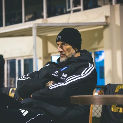 Na zdjęciu widoczny jest mężczyzna w czarnej kurtce i czapce, siedzący na krześle z założonymi rękami, podczas wydarzenia sportowego.