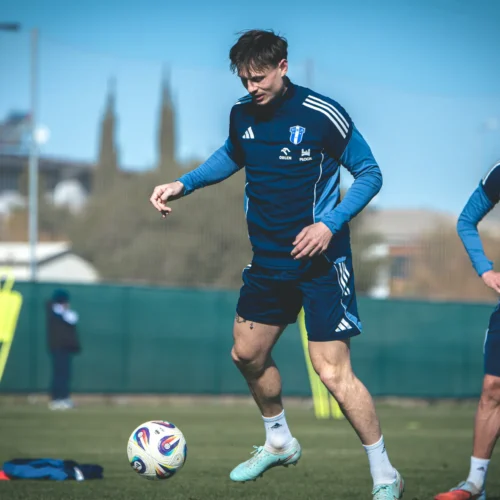 Na zdjęciu widać dwóch piłkarzy podczas treningu na boisku; jeden z zawodników wytrąca piłkę, a drugi obserwuje sytuację.