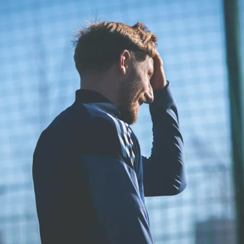 Mężczyzna w sportowym stroju przystaje na boisku, trzymając się za głowę w zamyśleniu.
